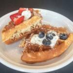 Close-up of two quarters of golden Belgian waffle on a white plate. One piece is topped with strawberries and whipped cream, and the other with blueberries and whipped cream. Both are dusted with powdered sugar.