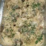 Overhead shot of a foil catering pan filled with Champagne Chicken, a dish covered in a thick, creamy pale gold sauce with sliced mushrooms and a generous topping of chopped parsley.