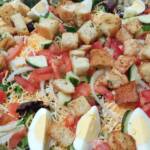 Close-up of a large, freshly prepared Sweet Leanne's Catering House Salad topped with herb croutons, sliced hard-boiled eggs, chopped tomatoes, cucumber slices, and shredded cheese over a bed of mixed greens and sliced onions.