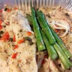 Close-up of baked fish fillets prepared for baking in a foil pan, covered with a lemon caper sauce, minced garlic, chopped red bell peppers, and black pepper. Four spears of fresh asparagus and slices of white onion sit in the center of the dish.