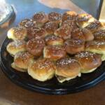 A large black round catering platter filled with numerous small, toasted ribeye sliders on sesame seed buns. The sliders are arranged in concentric circles on a dark brown surface.